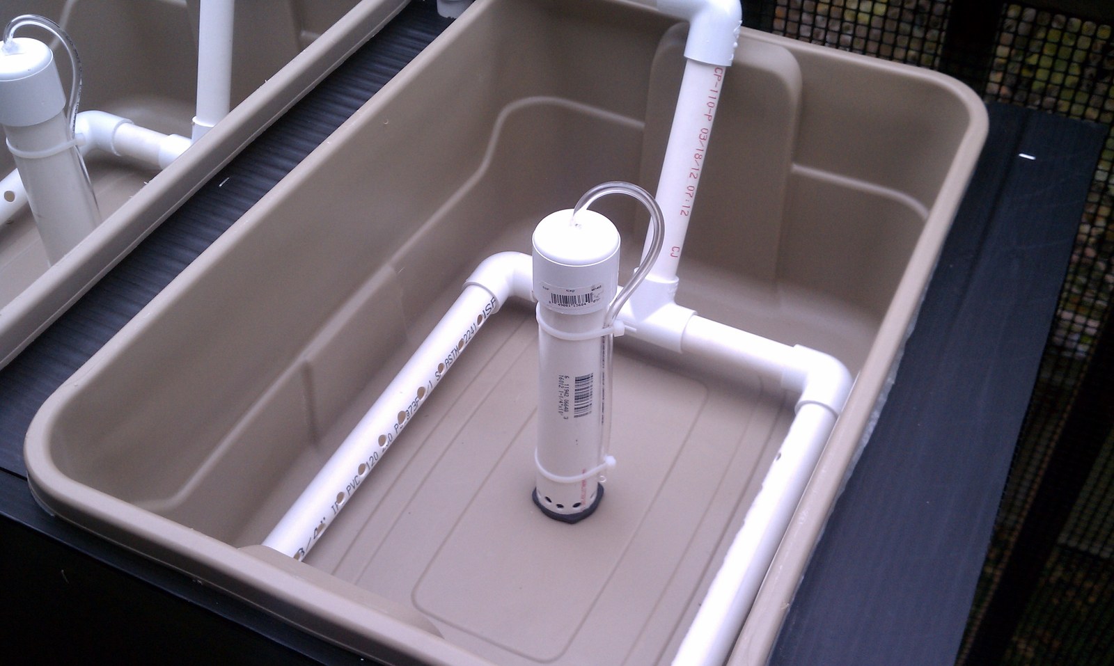 A shallow growing tub with a white PVC manifold and a black standpipe fitted, empty and dry.