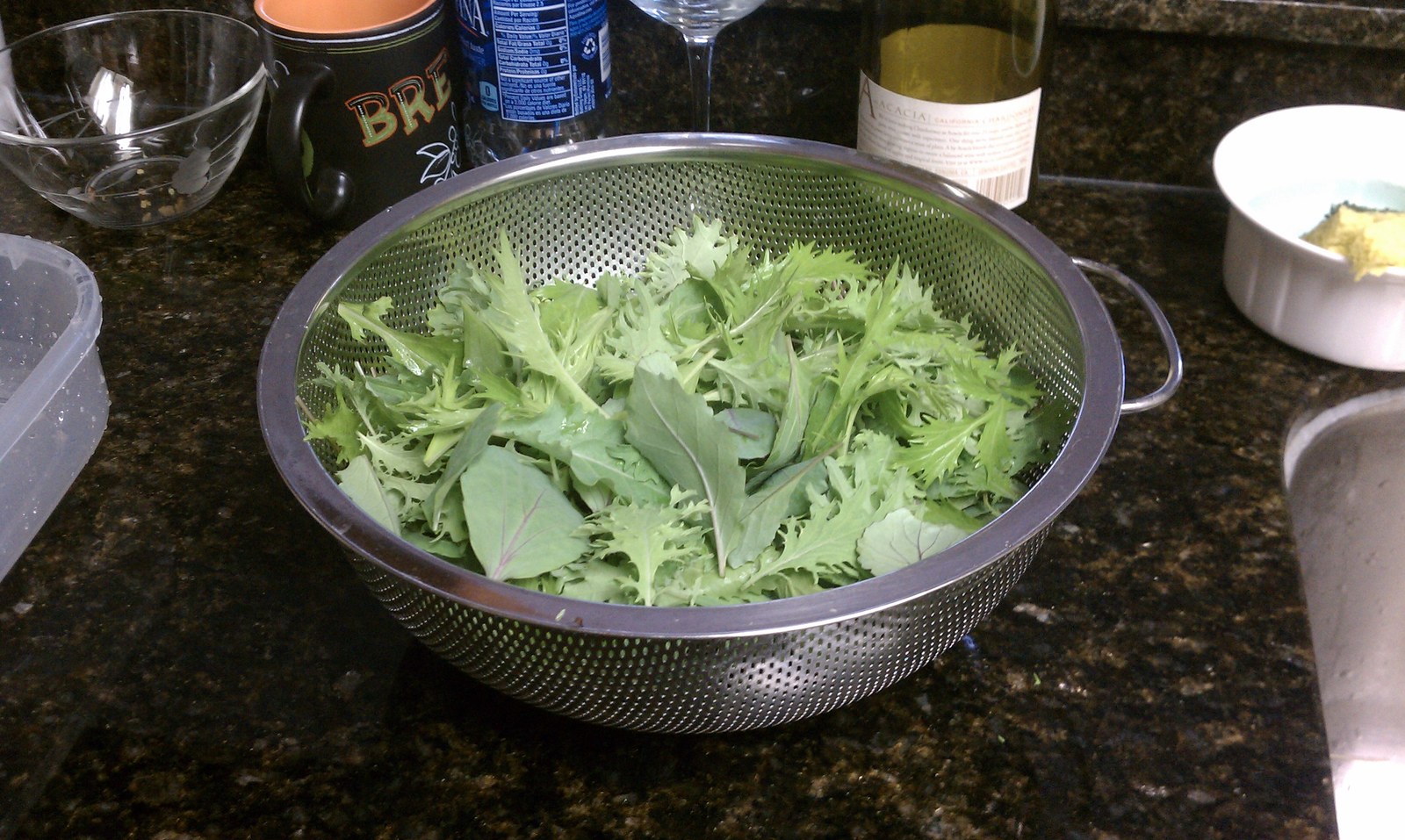 A steel colander heaped with freshly cut frilly greens on a dark granite kitchen counter.