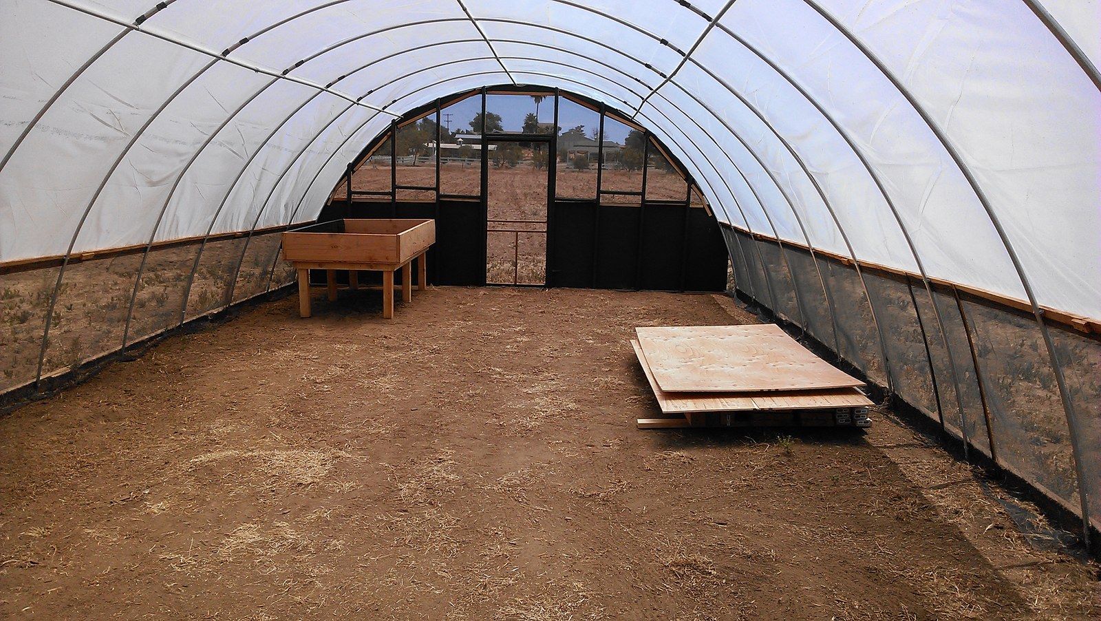 Inside the covered hoop house looking at the far end wall, bare earth floor, the first timber grow table built and plywood stacked on a pallet.