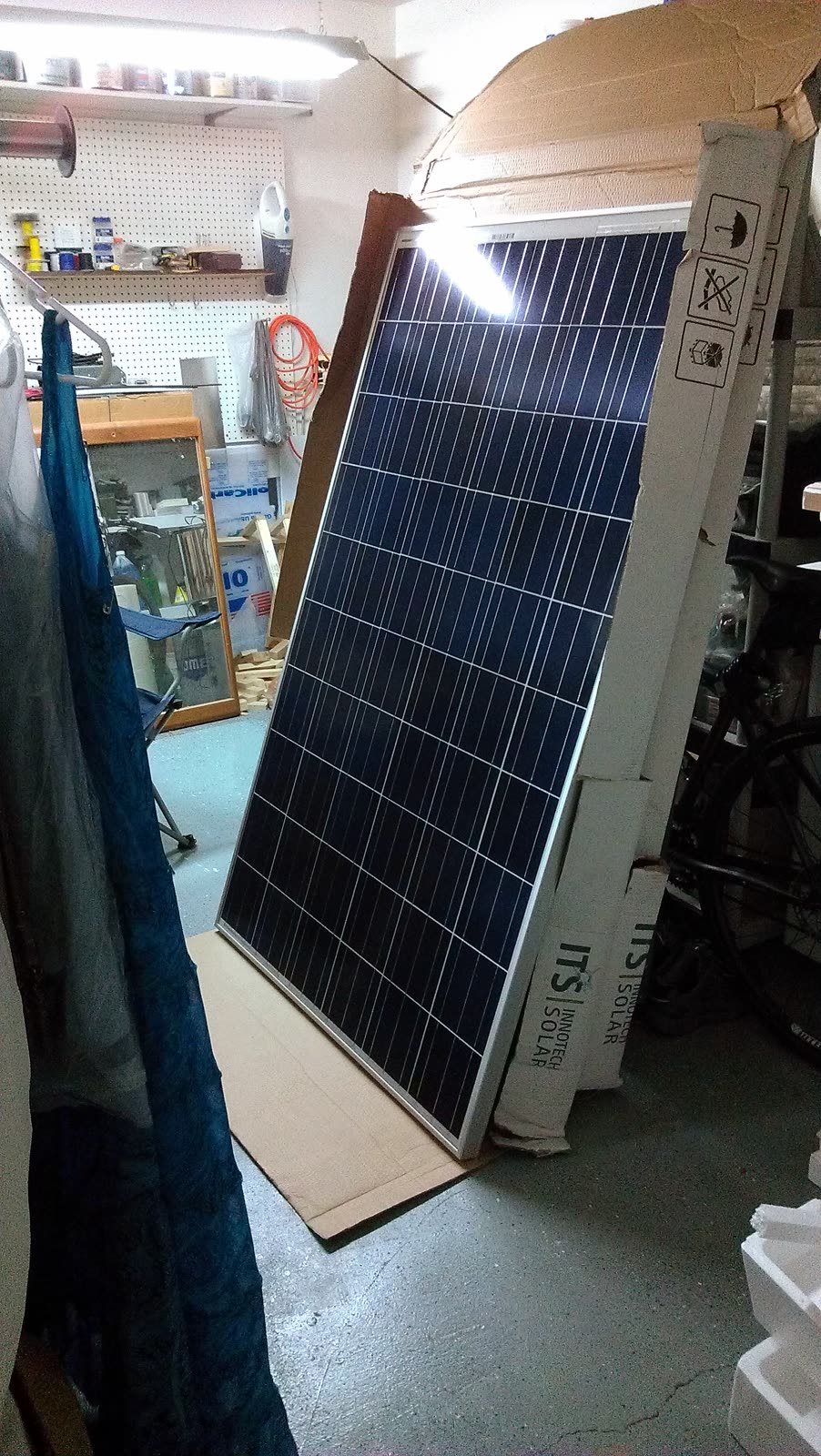 A large blue polycrystalline solar panel standing on end in a shop, still in its ITS Solar shipping wrap, next to the cardboard carton it came out of.