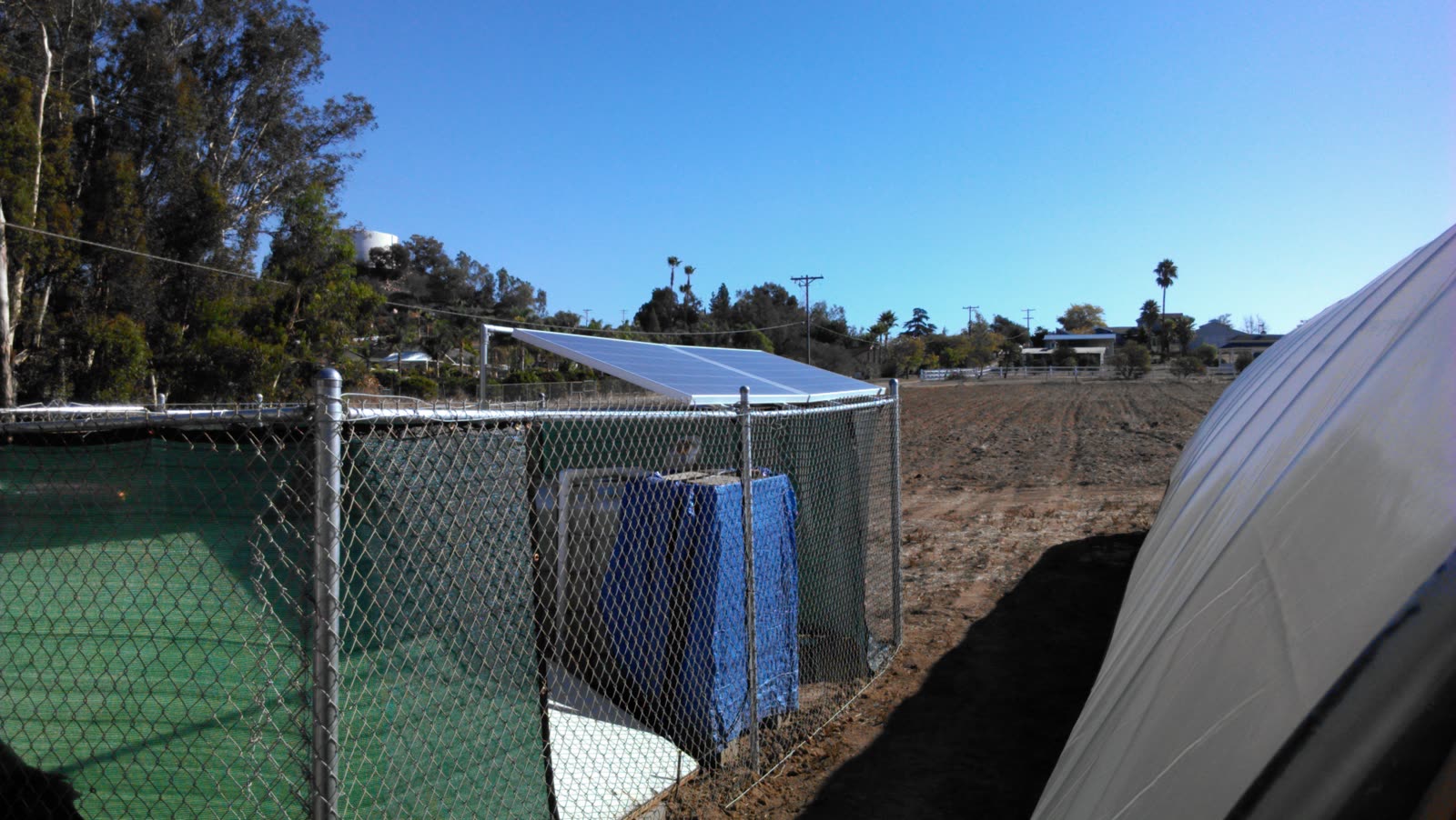 A solar panel on a tilt frame in full sun above a fenced enclosure, with the plowed field, the edge of the covered hoop house, and houses on the Fallbrook hillside behind it.