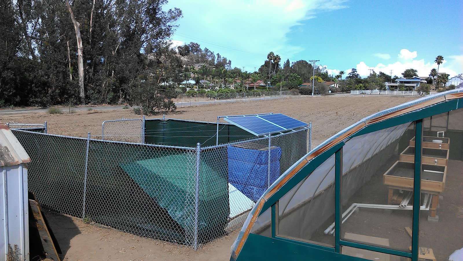 Two blue polycrystalline solar panels mounted on a steel tilt frame above a chain link pen at the farm, the greenhouse and its timber grow tables on the right, a plowed field and the Fallbrook hillside behind.