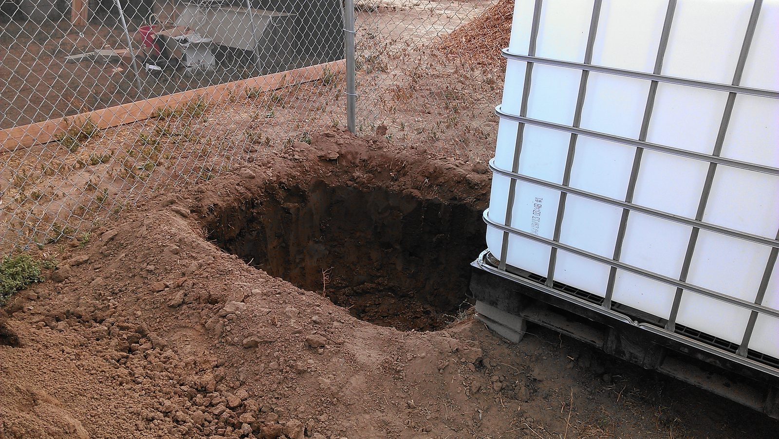 A pit dug in the red soil beside a white IBC tote on its pallet frame.