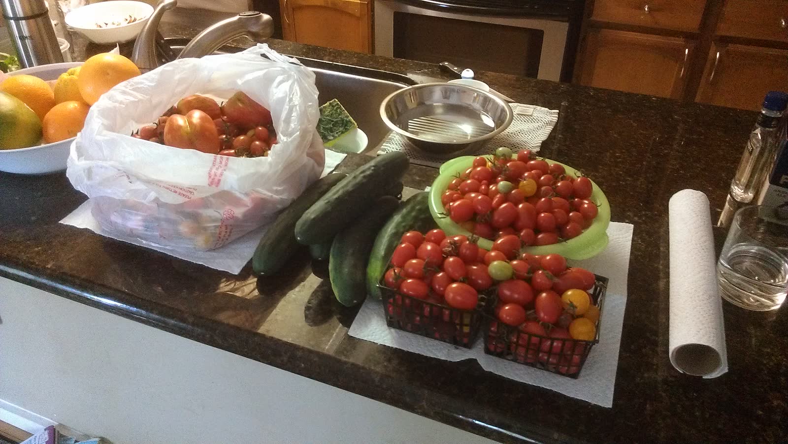A kitchen counter covered with one picking: whole cucumbers, two punnets of cherry tomatoes, larger tomatoes bagged, and a bowl of fruit behind.