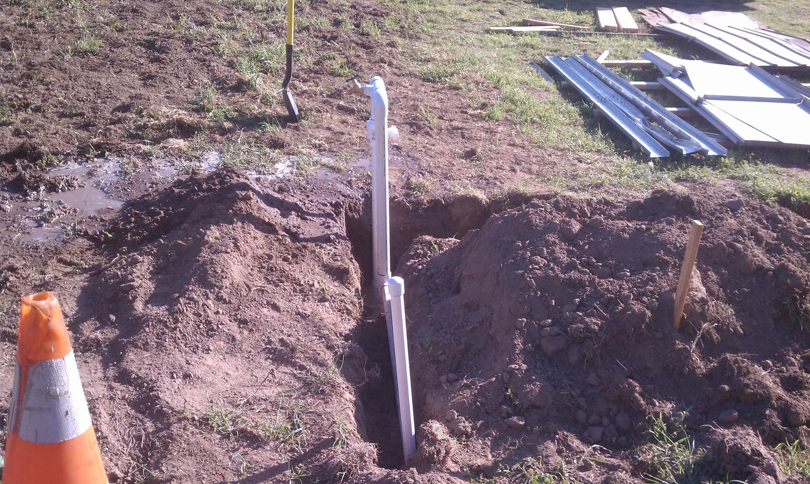 A white PVC riser standing in the partly backfilled trench with a shovel and cone nearby.