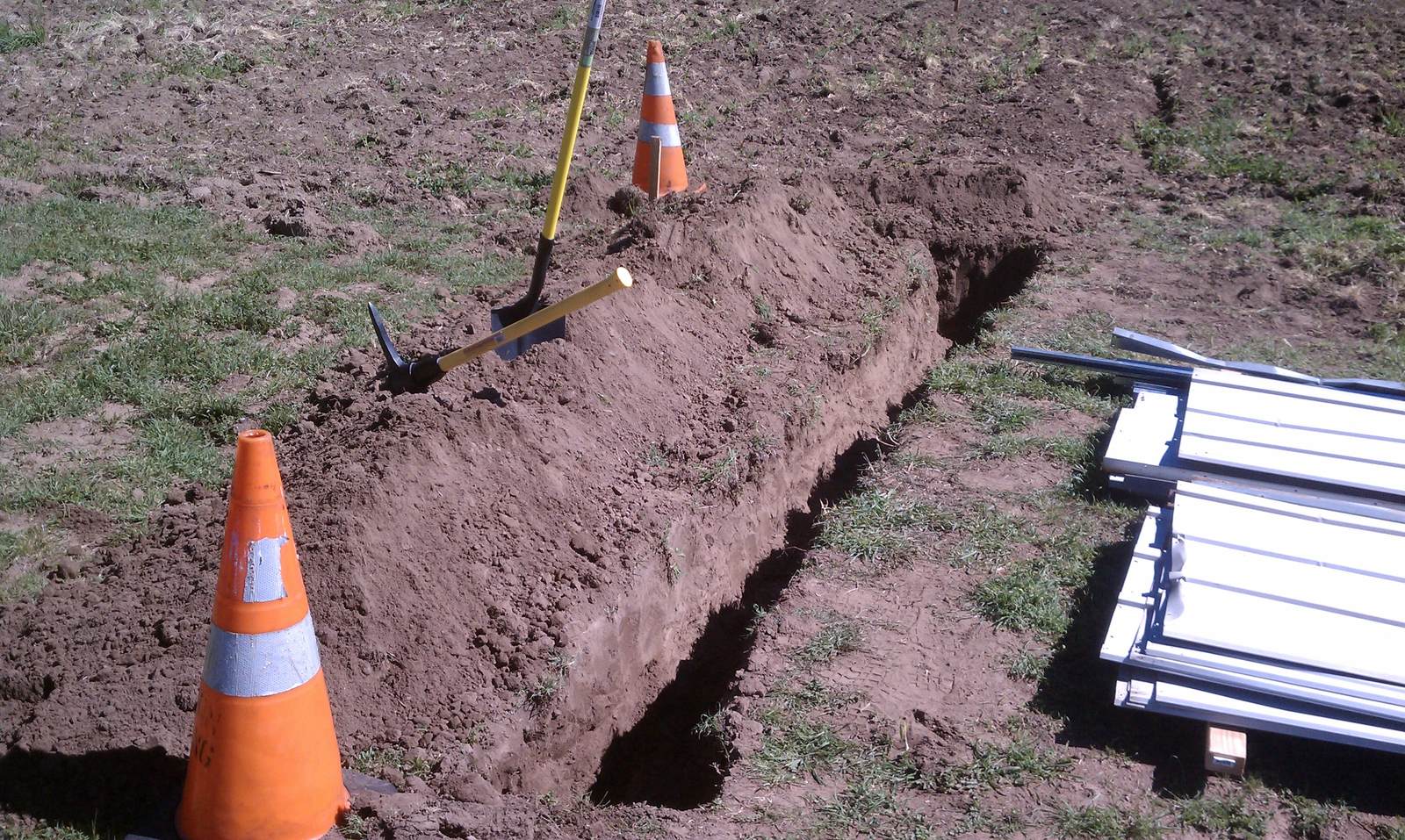 An open trench in the grass with a traffic cone, a pick, and stacked metal panels beside it.