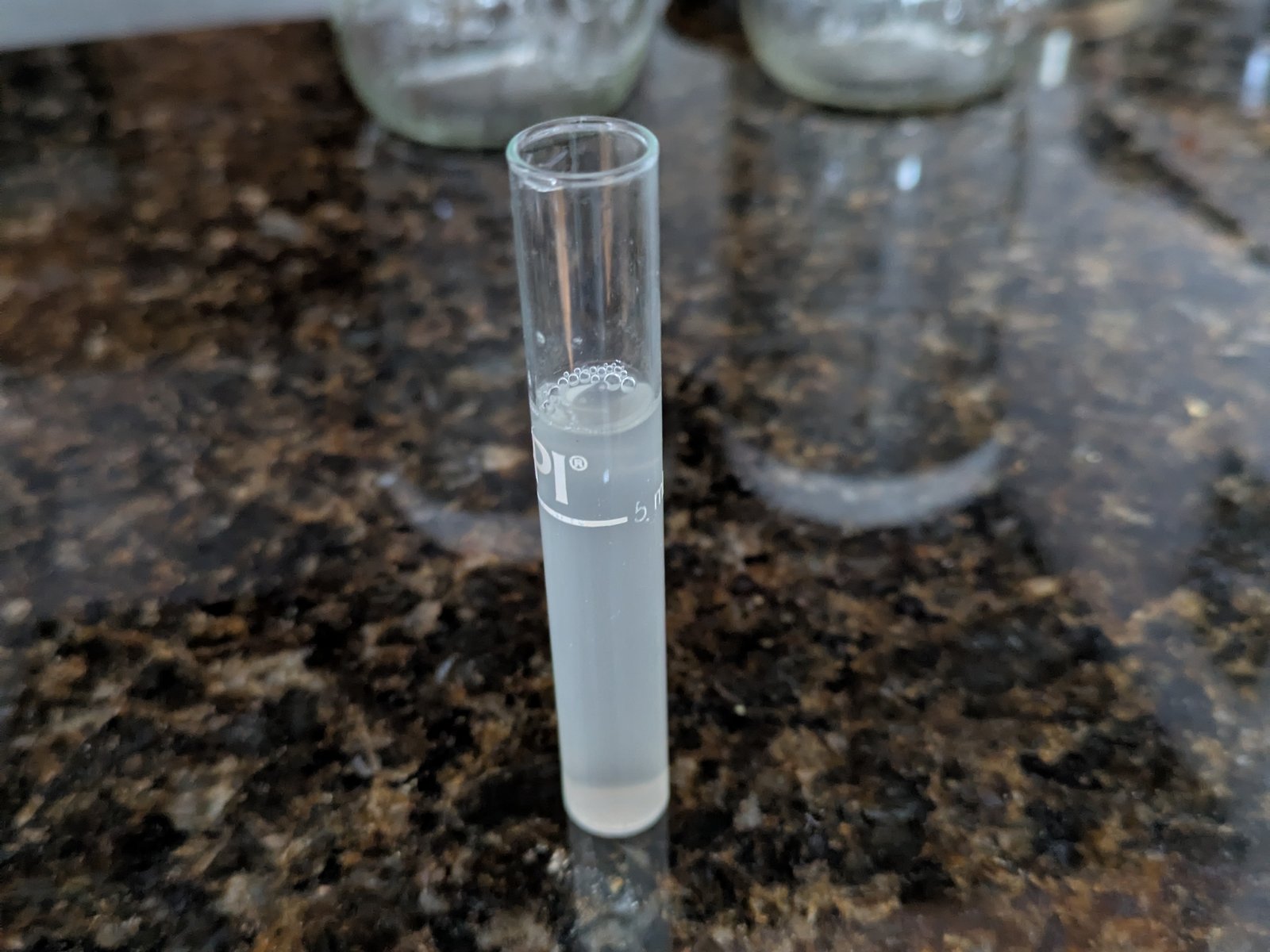 A five millilitre graduated sample tube holding drawn-off brine, standing on the counter with empty jars behind it.