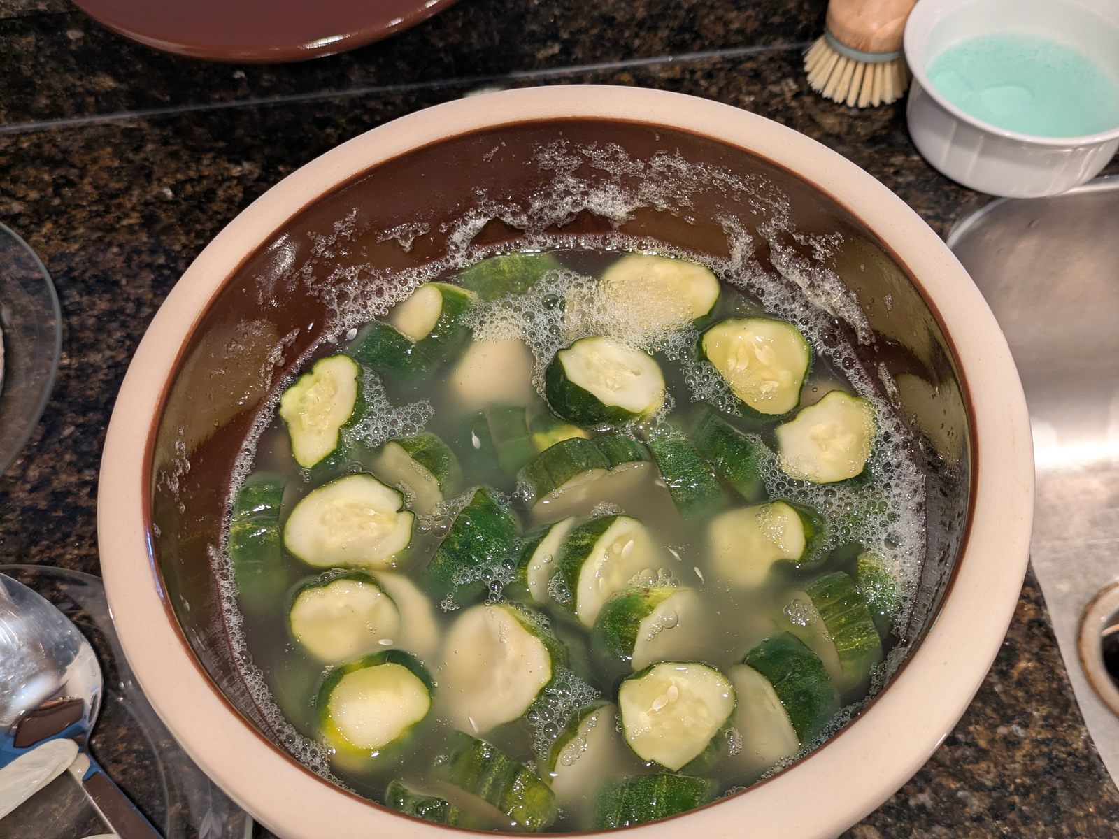 Cut cucumbers held under clear brine in a stoneware crock, a thick ring of fermentation bubbles standing all the way around the brine line.