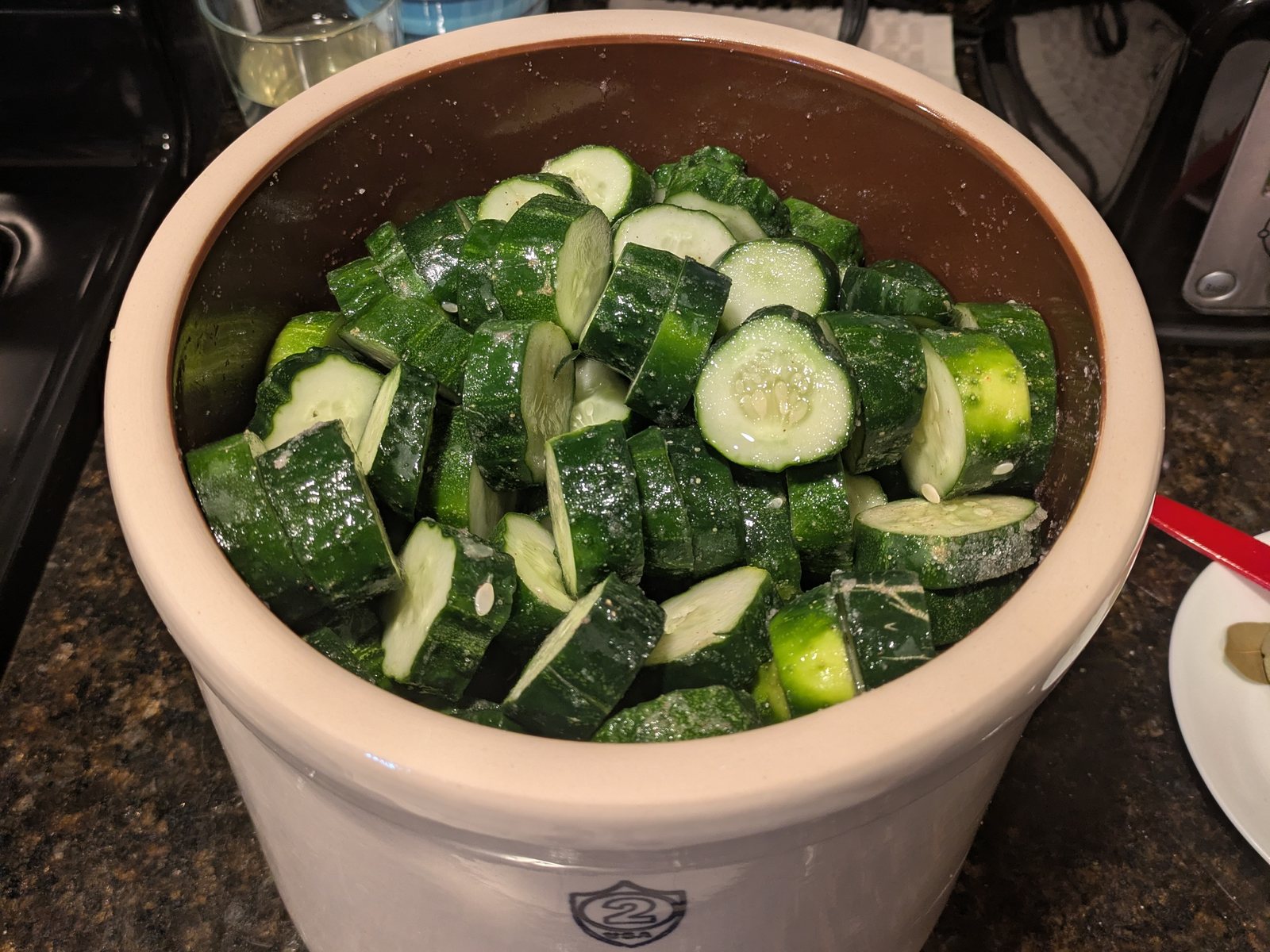 A two-gallon stoneware crock packed to the shoulder with cut cucumbers, salt still visible undissolved on the cut faces, a plate of bay leaves at the edge of the counter.