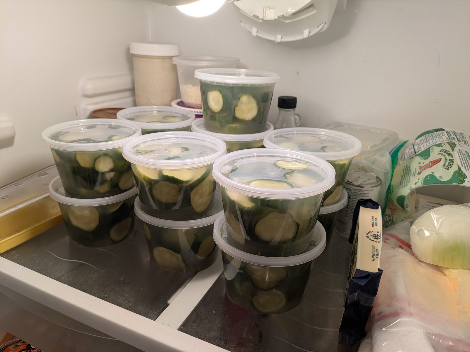 A refrigerator shelf stacked two deep with about ten clear deli containers of finished pickle chips, each one covered in brine.