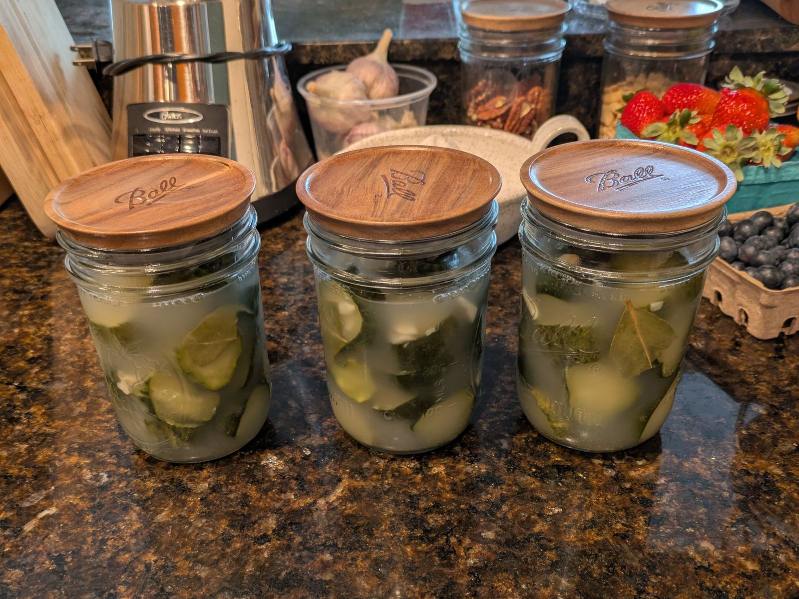 Three wide-mouth jars of finished cucumber chips under cloudy brine, whole bay leaves and garlic cloves visible through the glass, wooden lids on top.