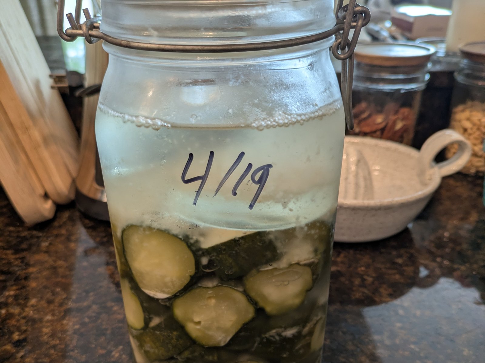 A clip-top jar of cucumber chips under brine with the date 4/19 written large on the glass in marker, bubbles standing at the surface.