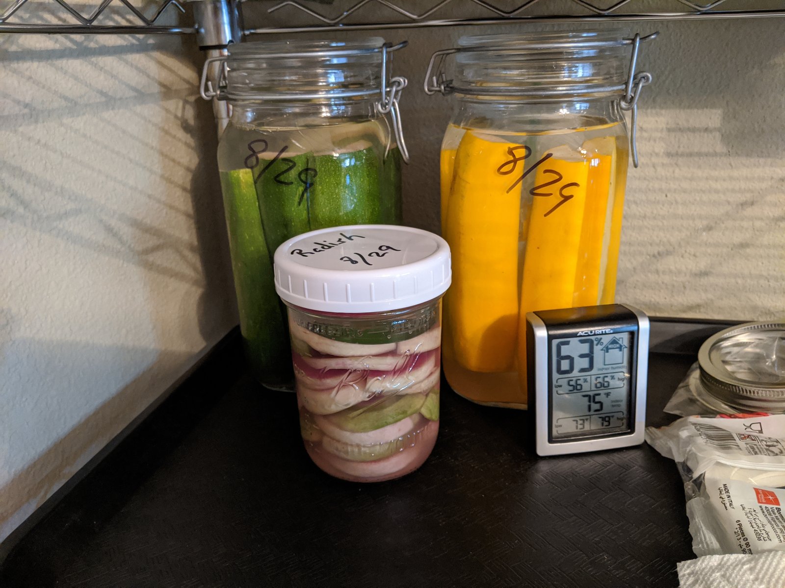 Two clip-top jars of whole cucumbers and yellow squash and a smaller jar of radish, each dated 8/29 in marker, on a wire shelf beside an AcuRite gauge reading 63 percent humidity and 75 degrees with low and high figures beneath.